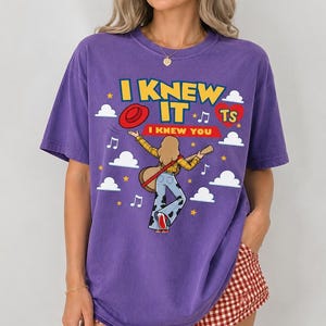 May include: A purple short-sleeve t-shirt featuring a graphic of a person with blonde hair playing a guitar, wearing a yellow plaid shirt and cow-print bell-bottom pants. The text reads I KNEW IT and I KNEW YOU, surrounded by clouds and stars.