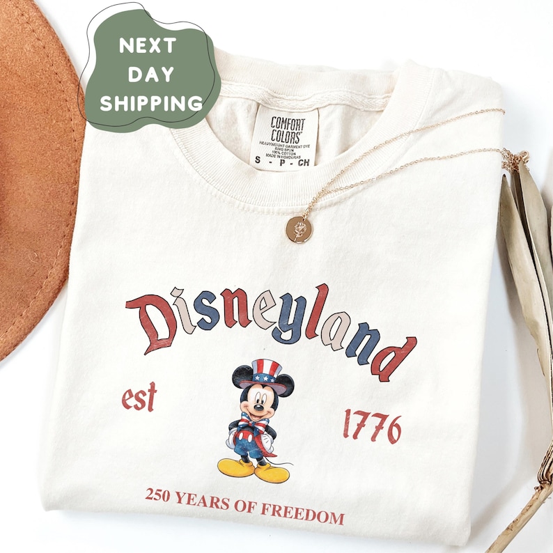 A white t-shirt with a Disneyland logo and text, along with a necklace and a brown hat, is displayed on a white background.