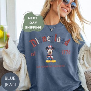 A woman wearing a blue t-shirt with a Mickey Mouse graphic and text, smiling and holding a drink.