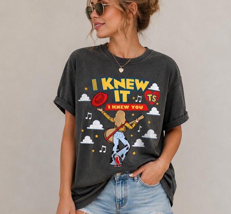 May include: A charcoal gray t-shirt featuring a graphic print with the text I KNEW IT I KNEW YOU. The design includes a blonde figure playing a guitar, a red hat, a red heart with TS, white clouds, musical notes, and yellow stars on the front.