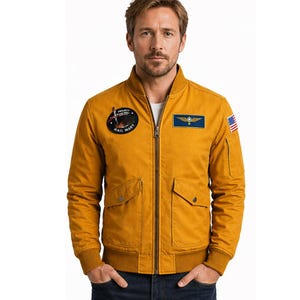 Puede incluir: Chaqueta bomber de color amarillo mostaza con cremallera, dos bolsillos delanteros y puños y dobladillo de canalé. La chaqueta presenta un parche circular con el texto "PROJECT HAIL MARY" y un parche rectangular con un diseño azul y dorado, además de una pequeña bandera estadounidense.