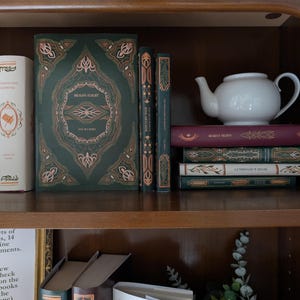 Può includere: Una collezione di libri con copertina rigida dai motivi decorati in verde, crema e bordeaux con dettagli in lamina d'oro, inclusi titoli come Dragon Flight, King Arthur e Arabian Nights, disposti su uno scaffale in legno con una teiera bianca.