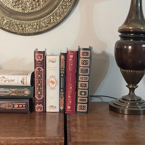 Pode incluir: Uma coleção de livros antigos com capas decorativas, dispostos sobre uma superfície de madeira. Os livros apresentam várias cores, incluindo bordô, creme e verde, com detalhes dourados ornamentados. Uma placa de latão e uma lâmpada vintage estão no fundo.