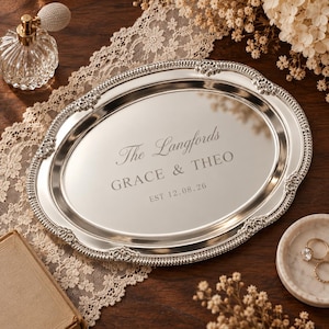 May include: An oval silver-toned metal tray with an ornate, beaded, and scrolled border. The centre is engraved with the text "The Langfords", "GRACE & THEO", and "EST 12.08.26" in a serif font. The tray has a polished, reflective surface.