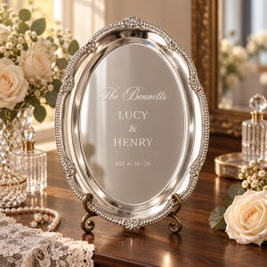 Pode incluir: Uma bandeja oval de cor prateada com uma borda ornamentada de contas e volutas, exibida num suporte de metal. O centro está gravado com o texto: The Bennetts, LUCY & HENRY, EST 6:18:26. A superfície refletora apresenta um acabamento liso e polido.