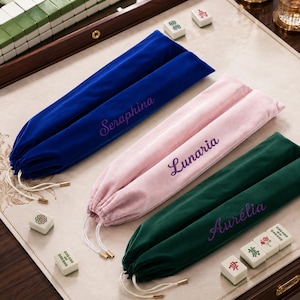 May include: Three long velvet drawstring pouches in royal blue, pale pink, and dark green. Each pouch features a name embroidered in purple script: Seraphina on the blue, Lunaria on the pink, and Aurélia on the green. Each bag has white cords.
