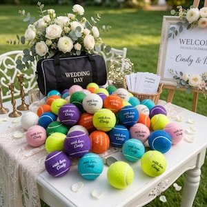 May include: A collection of multi-colored tennis balls scattered on a white table, each printed with the text 'with love, Cindy'. The balls are in shades of yellow, purple, orange, blue, green, pink, and white, arranged near a black bag.