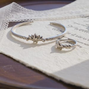 May include: A silver-toned bracelet and ring set. The bracelet is a cuff style with a floral design in the center and beaded accents. The matching ring features a similar floral motif. Both pieces are displayed on a textured, off-white surface.