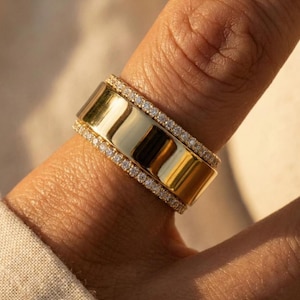 Puede incluir: Un anillo ancho de color dorado con una superficie pulida y reflectante. El anillo está adornado con una fila de pequeñas piedras brillantes a cada lado, creando una banda brillante. El anillo se lleva en un dedo, mostrando su diseño.