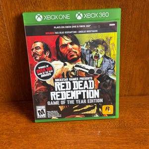 May include: A green Xbox One game case featuring the game "Red Dead Redemption: Game of the Year Edition." The cover art depicts three men and a zombie. The case includes the text "Includes Red Dead Redemption Undead Nightmare."