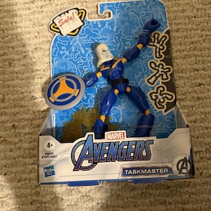 Peut inclure: Figurine Marvel Avengers Taskmaster dans un emballage bleu et blanc. La figurine est bleue avec des accents jaunes et un casque blanc. L'emballage porte le logo Avengers et le mot "Sale!" dans une bulle jaune.