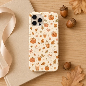 May include: A cream-colored phone case featuring a repeating pattern of orange pumpkins, white ghosts, autumn leaves, bows, and coffee drinks. The design includes small star accents and floral elements in shades of orange, brown, and tan.