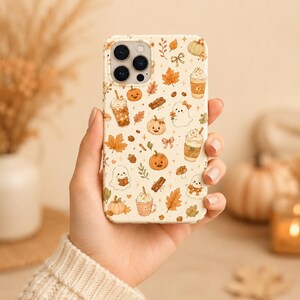 May include: A cream-colored smartphone case featuring a repeating pattern of autumn-themed illustrations including orange pumpkins, ghosts, cinnamon sticks, maple leaves, coffee cups with whipped cream, small bows, and tiny sparkling stars.