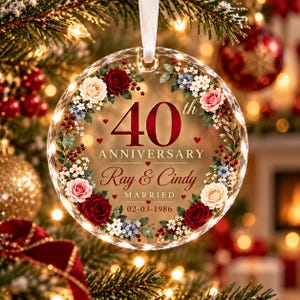 Op de afbeelding: Een rond, helder glazen ornament met een bloemenkrans van rode, roze en witte rozen met blauwe accenten. De tekst luidt 40th ANNIVERSARY, Ray & Cindy, MARRIED en 02-03-1986, versierd met kleine rode hartjes en een wit lint als hanger.