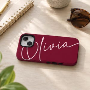 May include: A deep red smartphone case featuring the name Olivia written in a light pink cursive script across the back. The case includes a black camera lens cutout and black side buttons, designed to fit a dual-lens smartphone model.
