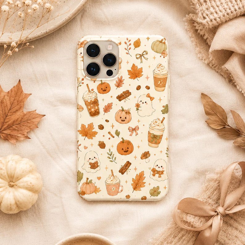 May include: A cream-colored phone case featuring a repeating pattern of autumn-themed illustrations including orange pumpkins, ghosts, cinnamon sticks, coffee cups with whipped cream, maple leaves, small bows, and tiny sparkling stars.