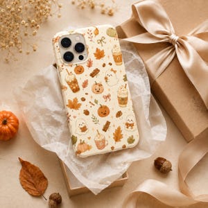 May include: A cream-colored smartphone case featuring a repeating pattern of autumn-themed illustrations including orange pumpkins, ghosts, cinnamon sticks, maple leaves, acorns, bows, and iced coffee drinks with whipped cream toppings.