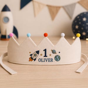 May include: A beige fabric crown featuring embroidered space-themed designs including a rocket, planets, stars, the number 1, and the name OLIVER. The crown is topped with five small colorful pom-poms in pink, light blue, orange, white, and yellow.
