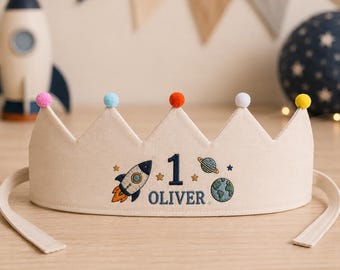 May include: A beige fabric crown featuring embroidered space-themed designs including a rocket, planets, stars, the number 1, and the name OLIVER. The crown is topped with five small colorful pom-poms in pink, light blue, orange, white, and yellow.