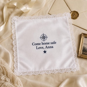 May include: A square white handkerchief with a delicate lace border. The center features dark blue embroidery of a compass rose, the text "Come home safe Love, Anna," and a small five-pointed star positioned below the text.