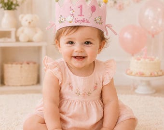 May include: A light pink felt crown featuring a white duck wearing a party hat, the number 1, a pink balloon, and the name Sophia embroidered in pink. The crown is topped with five small colorful pom-poms and has side ties for fastening.