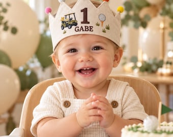 May include: A beige fabric crown featuring embroidered golf-themed graphics, including a golf cart, a golf ball on a tee, and the number 1 with the name GABE. The crown is topped with small, colorful pom-poms in pink, blue, orange, and yellow.