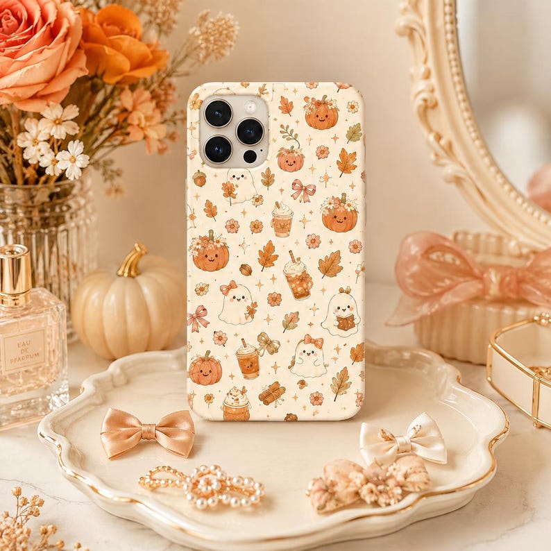 May include: A cream-colored phone case featuring a repeating pattern of orange pumpkins, white ghosts, autumn leaves, bows, and iced coffee drinks. The design includes small star accents scattered across the surface, with a camera cutout at the top.
