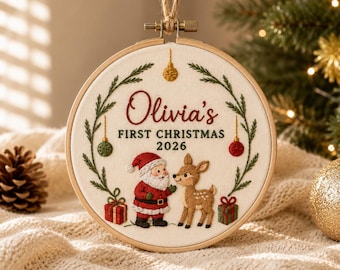 May include: A round wooden embroidery hoop containing off-white fabric with hand-stitched embroidery. The text reads Olivia's FIRST CHRISTMAS 2026. It features a Santa Claus figure, a small deer, two gift boxes, and a green pine branch wreath.