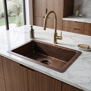Puede incluir: Un fregadero de cocina rectangular de cobre martillado con acabado marrón oscuro, que incluye un desagüe a juego, combinado con un grifo alto y arqueado de oro cepillado. El fregadero está integrado en una encimera de mármol blanco con vetas grises.