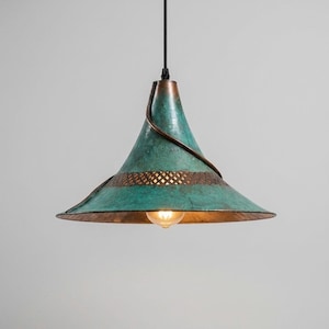 Può includere: Una lampada a sospensione conica con una finitura patinata color turchese e rame. Il paralume presenta una fascia decorativa in rete metallica marrone scuro e un dettaglio a filo a spirale, sospesa da un sottile cavo nero con una lampadina.