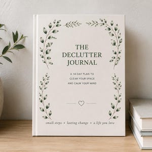 Op de afbeelding: Een hardcover journal met de titel "The Declutter Journal" met een crèmekleurige kaft. De kaft heeft een botanisch ontwerp en de tekst "A 14-day plan to clear your space and calm your mind". Er staat een klein hartje onder de tekst.