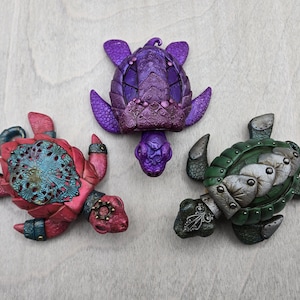 Peut inclure: Trois figurines décoratives de tortues dans les tons de rouge, violet et vert. Les tortues ont des détails complexes, notamment des carapaces texturées et des accents décoratifs. Les figurines sont exposées sur une surface claire.