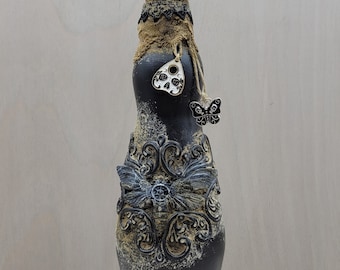 May include: A black glass bottle decorated with a skull, butterflies, and a Ouija board charm. The bottle is covered in a distressed, aged finish with a burlap-like texture.