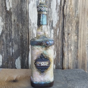 Peut inclure: Une bouteille en verre décorative avec un bouchon en cristal. La bouteille est peinte avec un aspect vieilli et vintage et présente une étiquette en métal avec des engrenages et une bordure en dentelle.