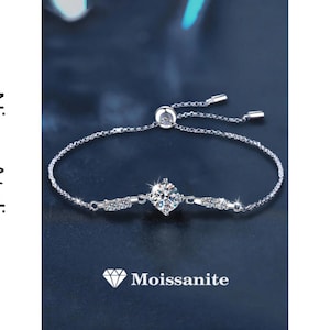 May include: A delicate silver bracelet with a central, sparkling moissanite gemstone. The bracelet features a sliding clasp for adjustable sizing and is adorned with smaller accent stones. The image includes the text "Nivra Atelier" and "Moissanite".