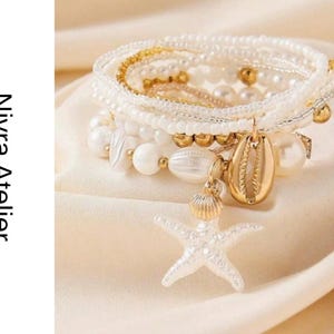 May include: A multi-strand bracelet set featuring white beads, pearls, and gold-toned accents. The set includes a gold-toned cowrie shell charm and a white starfish charm. The text Nivra Atelier is visible on the left side of the image.