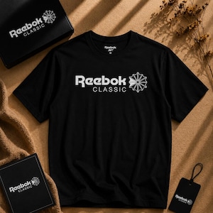 Op de afbeelding: Zwart Reebok Classic T-shirt met wit logo en tekst. Het logo heeft een gestileerd sterrenpatroon. Het shirt wordt gepresenteerd met een bijpassende doos, een label en een kleine takje gedroogde bloemen.