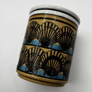 May include: A cylindrical ceramic mug with a tan background and black seashell designs. The mug has a white rim with black stripes and a blue band. The mug is approximately 4 inches tall.