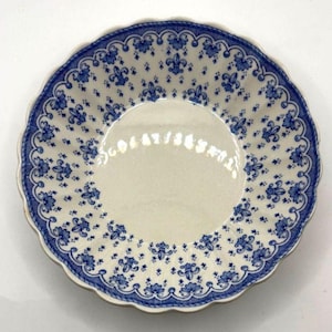 Könnte beinhalten: Eine runde Keramikschale mit gewelltem Rand, die eine cremefarbene Basis mit einem wiederkehrenden blauen Muster aus Fleur-de-Lis und kleinen Blumenmotiven aufweist. Der Rand ist mit einer durchgehenden blauen Bogenkante verziert.