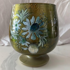 Peut inclure: Un verre à pied arrondi avec une finition vert olive métallique, orné d'un motif floral peint à la main dans des tons blancs et bleus. L'illustration comprend des pétales détaillés, des feuilles et de petites taches dorées. Le nom Sasha est signé dans le coin inférieur droit.