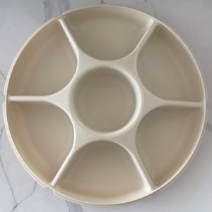 May include: A beige, round serving tray with six compartments and a central dip section. The tray is made of a smooth, solid material and is designed for serving appetizers, snacks, or dips. The compartments are separated by raised dividers.