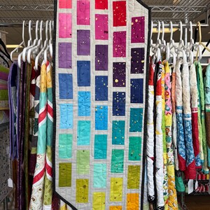 May include: A colorful, handmade quilt with a gradient of rectangular blocks in shades of red, pink, purple, blue, green, and yellow. The blocks are set against a light gray background and accented with gold flecks. The quilt is displayed on a rack with other quilts.