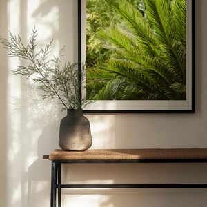 Op de afbeelding: Een vierkante ingelijste wandprint met een levendig groen palmbladontwerp, geplaatst boven een zwarte metalen consoletafel met een geweven blad. Een donkergrijze, getextureerde keramische vaas met een groene tak staat op de tafel.