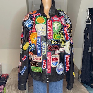 May include: Black leather jacket adorned with numerous colorful patches featuring sports team logos and names. The jacket has a high collar and a button-up front, showcasing a vintage aesthetic. Patches include "Lakers," "Capitals," and "Pacers."
