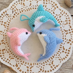 May include: Three small crocheted dolphin toys in light blue, pink, and medium blue. Each dolphin features a white underbelly, a curved dorsal fin, side fins, and a small black bead eye. The toys are arranged in a circular formation on a white lace doily.