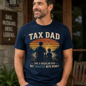 May include: Navy blue t-shirt with the text "TAX DAD" above a graphic of a sunset and two figures. Below the graphic, the text reads "LIKE A REGULAR DAD BUT SMARTER WITH MONEY". The shirt is a casual wear item.