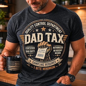 Op de afbeelding: Donkergrijs t-shirt met een "DAD TAX" ontwerp. Het ontwerp bevat de tekst "QUALITY CONTROL DEPARTMENT", "CERTIFIED DAD DUTY" en "SAMPLING REQUIRED - 1 BITE MINIMUM". Een zwarte mok met een Amerikaanse vlag is ook zichtbaar.