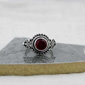 May include: A silver ring featuring a round, deep red gemstone set in the center. The setting is surrounded by a twisted rope texture and intricate, symmetrical scrollwork patterns on both sides of the band, creating a detailed metal design.