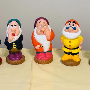 May include: A set of seven colourful figurines depicting characters with distinct facial expressions, hats, and outfits, each standing on a circular brown base. The figures feature varied colour schemes including blue, green, yellow, orange, and purple accents.
