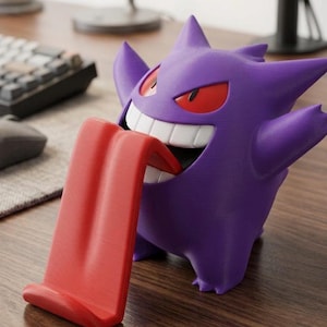 May include: A purple Gengar character figure serving as a phone or device stand. The figure features a wide, grinning mouth with white teeth and a long, protruding red tongue that acts as a support ledge for holding a mobile device upright.